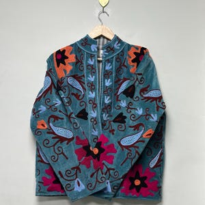 May include: Teal velvet jacket with detailed floral embroidery in pink, orange, blue, and black. The jacket has a high collar and a button-up front. The embroidery includes paisley and floral designs, creating a bohemian aesthetic.