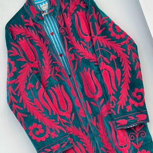 May include: A long-sleeved teal coat featuring vibrant pink floral embroidery throughout. The garment has a collar, a button-down front with small red heart-shaped buttons, and a light blue and white vertical striped lining visible at the neck.