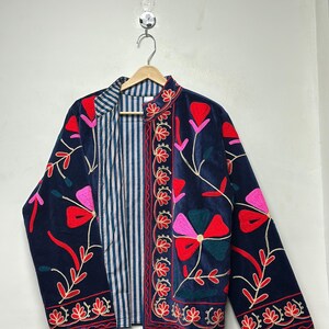Op de afbeelding: Een donkerblauw fluwelen jasje met levendige bloemenborduursels in rood, roze en groen. Het jasje heeft een opstaande kraag, lange mouwen en een open voorzijde die een blauw-witte voering met verticale strepen aan de binnenzijde toont.