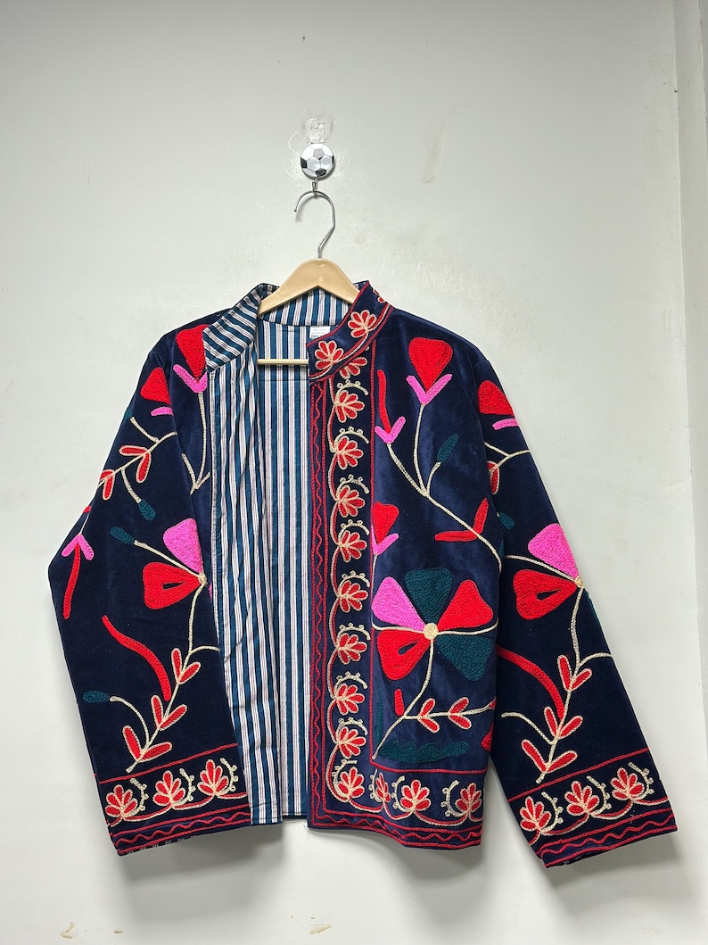 Op de afbeelding: Een donkerblauw fluwelen jasje met levendige bloemenborduursels in rood, roze en groen. Het jasje heeft een opstaande kraag, lange mouwen en een open voorzijde die een blauw-witte voering met verticale strepen aan de binnenzijde toont.