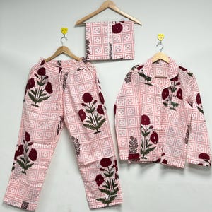 May include: A light pink pajama set with a floral design. The set includes a button-down shirt, trousers, and a folded scarf. The floral design features dark red flowers and green leaves. The fabric has a geometric grid pattern.