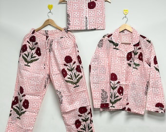 Puede incluir: Un conjunto de pijama rosa claro con un estampado floral. El conjunto incluye una camisa con botones, pantalones y una bufanda doblada. El diseño floral presenta flores de color rojo oscuro y hojas verdes. La tela tiene un patrón de cuadrícula geométrica.