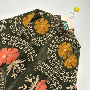 May include: Olive green velvet jacket with intricate floral embroidery in gold and orange. The jacket features a high collar and is displayed on a wooden hanger. The embroidery includes flowers and swirling vines.