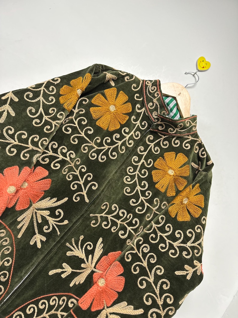 May include: Olive green velvet jacket with intricate floral embroidery in gold and orange. The jacket features a high collar and is displayed on a wooden hanger. The embroidery includes flowers and swirling vines.