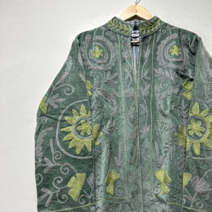 May include: A green velvet jacket with intricate floral embroidery in shades of gray and chartreuse. The jacket features a high collar and a full-length zipper. The design includes stylized flowers and leaf patterns.