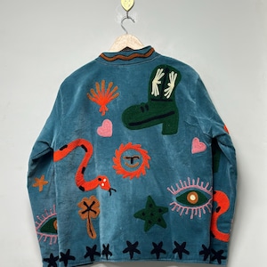 May include: Teal jacket with embroidered designs. Features a green boot, orange snake, and various other colorful embroidered shapes. The jacket is hanging on a wooden hanger against a light gray background.