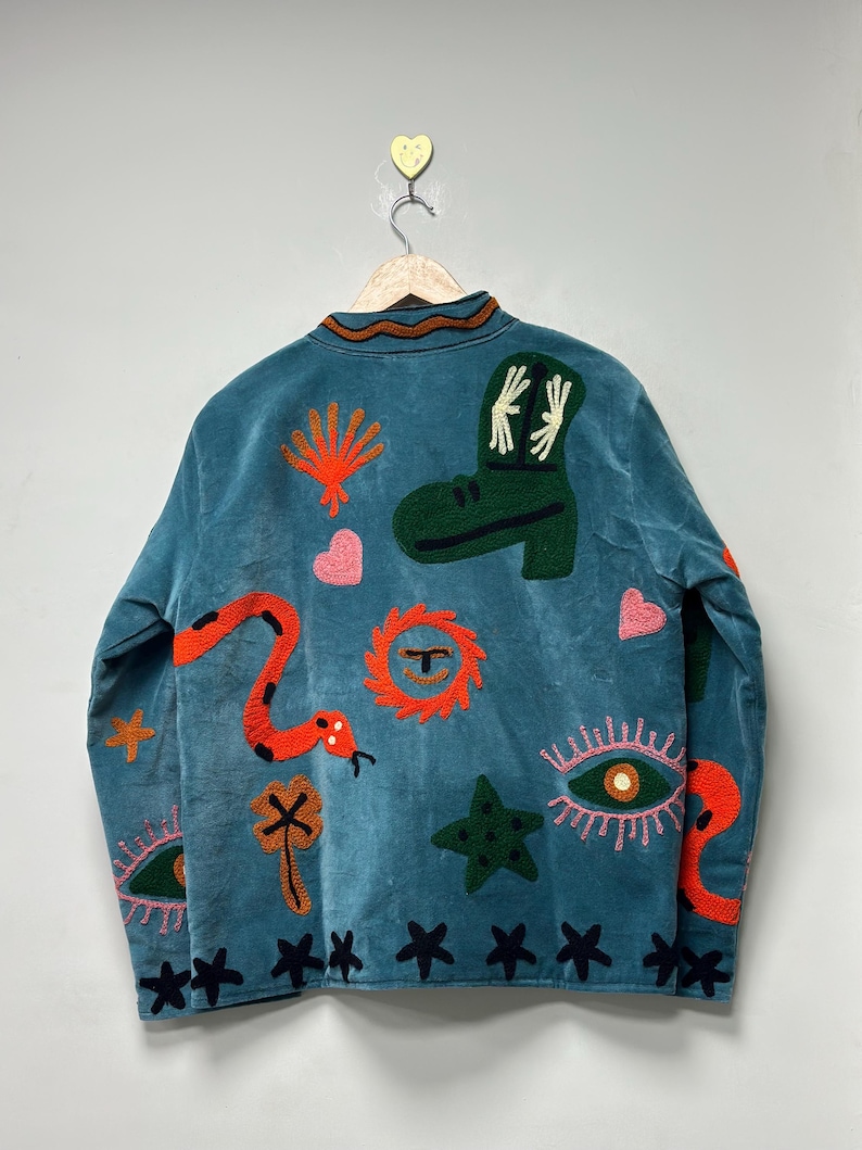 May include: Teal jacket with embroidered designs. Features a green boot, orange snake, and various other colorful embroidered shapes. The jacket is hanging on a wooden hanger against a light gray background.