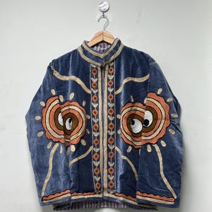 May include: A blue velvet jacket with elaborate embroidery. The jacket features large, circular designs in shades of orange, brown, and white, and gold-colored trim. The jacket is hanging on a wooden hanger.