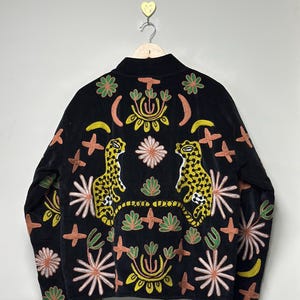 Może przedstawiać: Czarna kurtka z haftowanymi detalami. Kurtka ma wysoki kołnierz i jest ozdobiona kolorowymi wzorami kwiatowymi i zwierzęcymi. Tył kurtki przedstawia dwie żółto-czarne postacie przypominające lamparty, otoczone różnymi haftowanymi elementami.