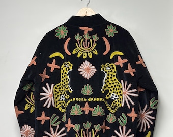 Puede incluir: Una chaqueta negra con bordados coloridos de dos leopardos amarillos, motivos florales, lunas crecientes y formas de cruz. El diseño es simétrico, con los leopardos enfrentados en el centro, rodeados por diversos patrones botánicos.