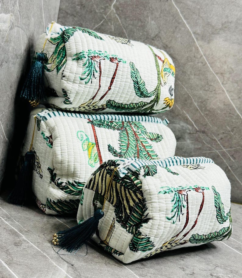 May include: Three white quilted zippered pouches with a tropical print featuring palm trees, leaves, and animals. The pouches have green and blue trim and tassels.