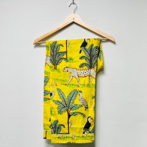 Puede incluir: Pantalones amarillos con estampado de tem&aacute;tica selv&aacute;tica. El dise&ntilde;o presenta un leopardo, tucanes, palmeras y otros elementos tropicales. Los pantalones est&aacute;n colgados en una percha de madera contra una pared blanca.