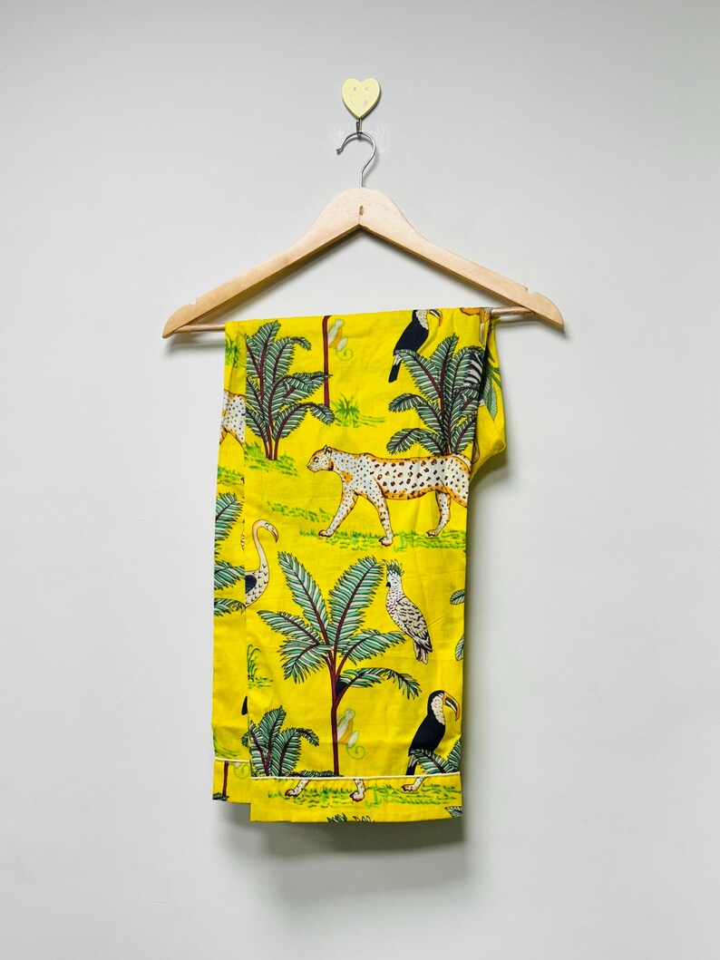 Puede incluir: Pantalones amarillos con estampado de tem&aacute;tica selv&aacute;tica. El dise&ntilde;o presenta un leopardo, tucanes, palmeras y otros elementos tropicales. Los pantalones est&aacute;n colgados en una percha de madera contra una pared blanca.