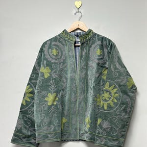 May include: A sage green velvet jacket with floral embroidery in shades of gray and chartreuse. The jacket has a mandarin collar and is hanging on a wooden hanger. The lining is a blue and white striped fabric.
