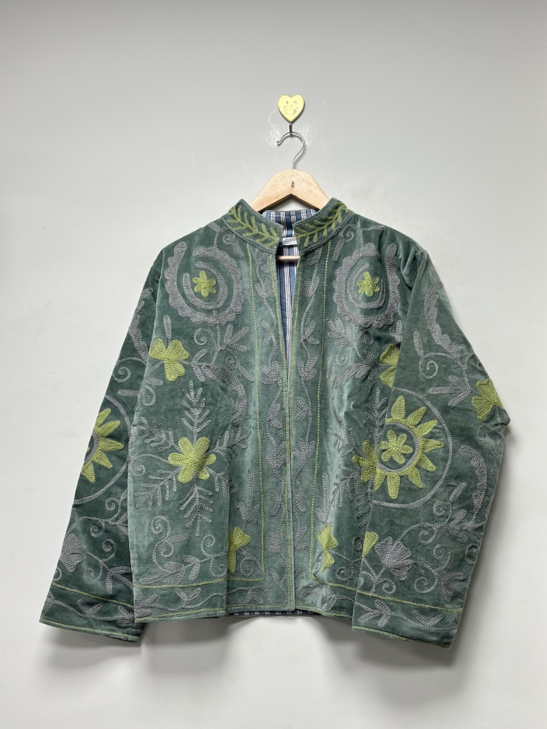 May include: A sage green velvet jacket with floral embroidery in shades of gray and chartreuse. The jacket has a mandarin collar and is hanging on a wooden hanger. The lining is a blue and white striped fabric.