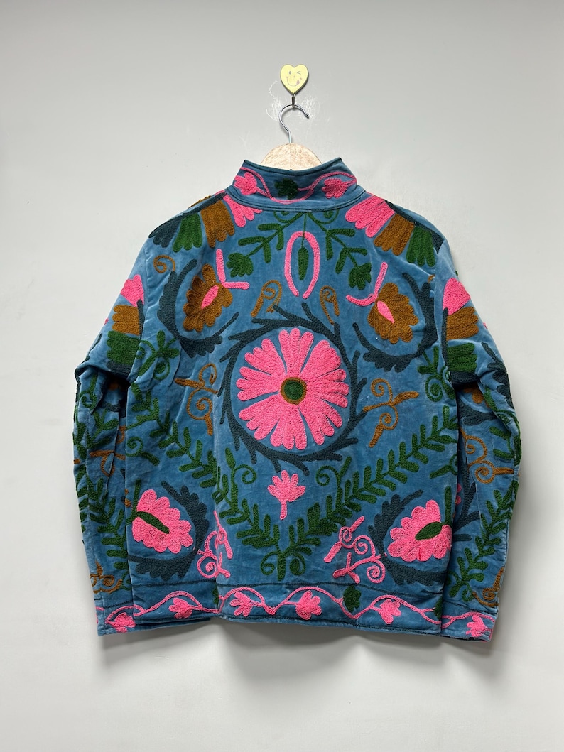 May include: A blue jacket with a high collar and intricate floral embroidery in pink, green, and brown. The jacket is hanging on a wooden hanger. The floral design is centered on the back.