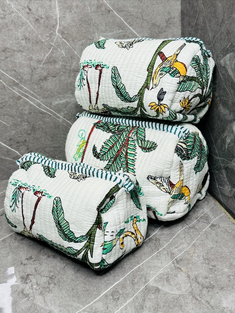 May include: Three white quilted fabric pouches with a green and white tropical print. The pouches are stacked on top of each other, with the largest pouch on the bottom and the smallest pouch on top. The pouches have a blue and white striped trim.
