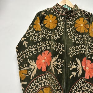 May include: Dark green velvet jacket with intricate floral embroidery in shades of gold, orange, and cream. The jacket features a high collar and a full-length zipper. The floral design includes large flowers and swirling vines, creating a detailed and ornate look.