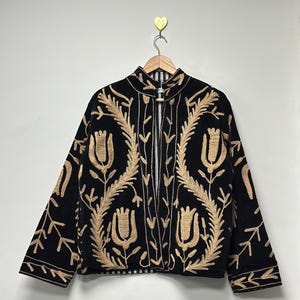 May include: A black jacket featuring long sleeves, a high collar, and an open front design. The garment is decorated with intricate tan-coloured embroidery depicting floral and leaf patterns across the front and sleeves. It hangs on a wooden hanger.