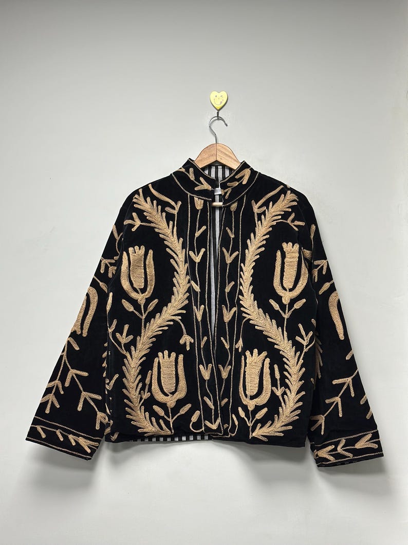 May include: A black jacket featuring long sleeves, a high collar, and an open front design. The garment is decorated with intricate tan-coloured embroidery depicting floral and leaf patterns across the front and sleeves. It hangs on a wooden hanger.
