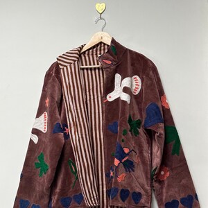 May include: A brown velvet jacket with a striped interior and colorful embroidered details. The jacket features appliqued birds, flowers, hearts, and leaves. The jacket is hanging on a wooden hanger.