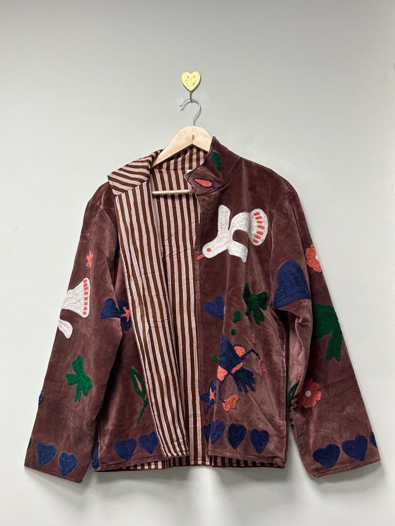 May include: A brown velvet jacket with a striped interior and colorful embroidered details. The jacket features appliqued birds, flowers, hearts, and leaves. The jacket is hanging on a wooden hanger.
