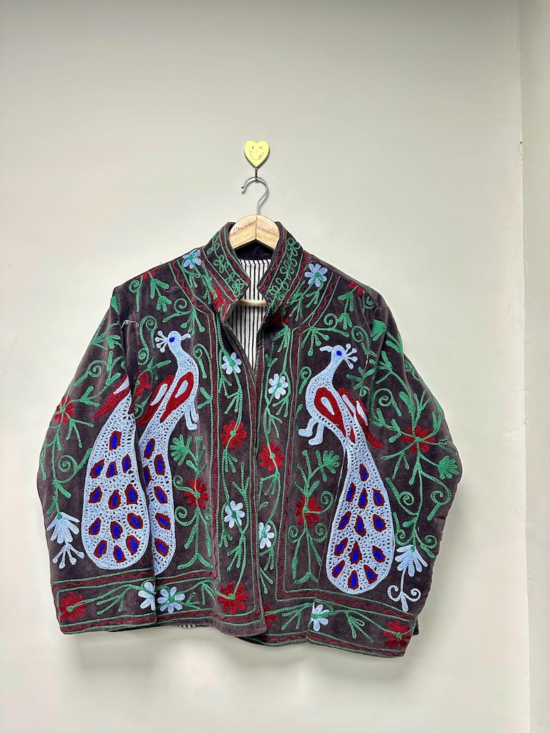 May include: A dark brown velvet jacket with embroidered peacock and floral designs. The jacket features a high collar and a striped lining. The peacock embroidery includes red, blue, and green accents, creating a vibrant and detailed pattern.
