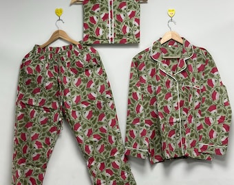 Puede incluir: Un conjunto de pijama verde con un estampado floral de flores rojas y blancas y hojas verdes. El conjunto incluye una camisa abotonada de manga larga, pantalones y una máscara para dormir plegada, todo con ribetes blancos. Los pijamas están colgados en perchas de madera.