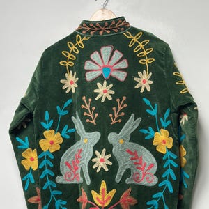 May include: A dark green velvet jacket featuring intricate embroidery of two rabbits, various flowers, and leafy stems in shades of yellow, light blue, pink, and cream. The jacket has a high collar and long sleeves with matching floral embroidery.