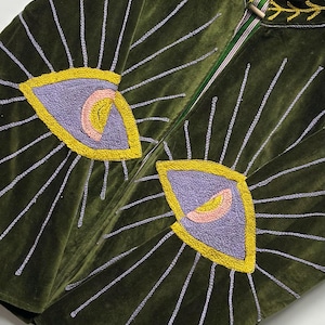 K&ouml;nnte beinhalten: Eine olivgr&uuml;ne Samtjacke mit gro&szlig;en gestickten Augenmotiven auf der Vorderseite. Die Augen sind mit violettem, gelbem und rosafarbenem Garn gestickt, mit hellvioletten Strahlen. Der Kragen ist mit einer gelben, blattartigen Stickerei versehen.