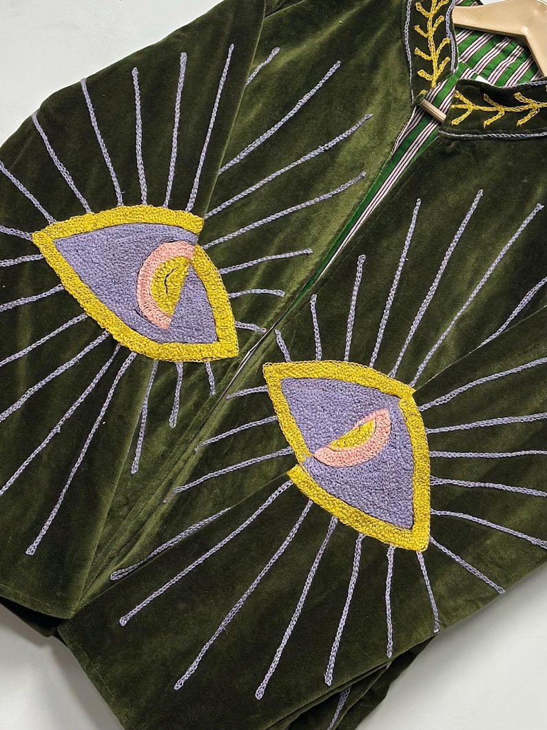 K&ouml;nnte beinhalten: Eine olivgr&uuml;ne Samtjacke mit gro&szlig;en gestickten Augenmotiven auf der Vorderseite. Die Augen sind mit violettem, gelbem und rosafarbenem Garn gestickt, mit hellvioletten Strahlen. Der Kragen ist mit einer gelben, blattartigen Stickerei versehen.