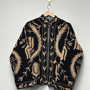 May include: A black velvet jacket featuring intricate gold-coloured embroidery in floral and leaf patterns. The jacket has a high collar, long sleeves, and a front closure with a metal toggle fastener. The interior lining is visible with black and white stripes.
