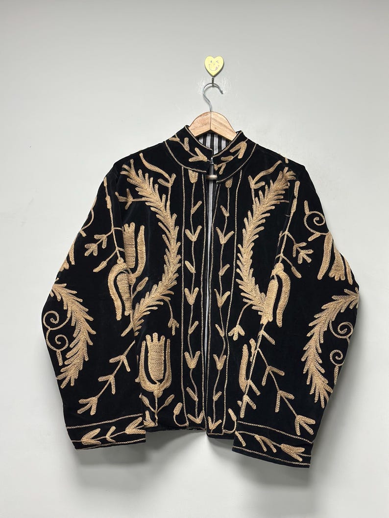 May include: A black velvet jacket featuring intricate gold-coloured embroidery in floral and leaf patterns. The jacket has a high collar, long sleeves, and a front closure with a metal toggle fastener. The interior lining is visible with black and white stripes.