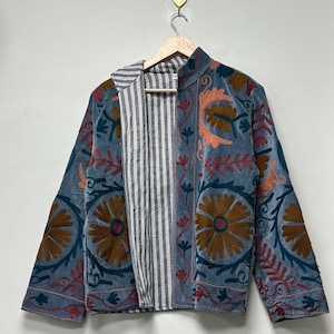 May include: A blue jacket with floral embroidery in shades of brown, orange, and red. The jacket has a striped lining and is hanging on a wooden hanger. The embroidery features large, stylized flowers and intricate leaf patterns.