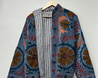 Puede incluir: Chaqueta azul con bordados florales en tonos marrón, naranja y rojo. La chaqueta tiene un forro a rayas y está colgada de una percha de madera. El bordado presenta grandes flores estilizadas y intrincados diseños de hojas.