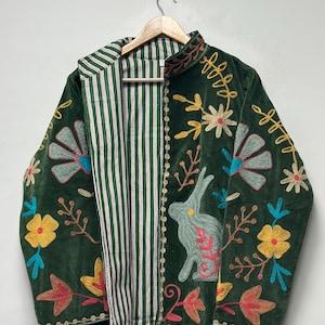 May include: A dark green velvet jacket featuring colorful embroidered floral patterns and a rabbit motif. The interior lining is made of white fabric with thin vertical green and red stripes. The jacket has long sleeves and a high collar.