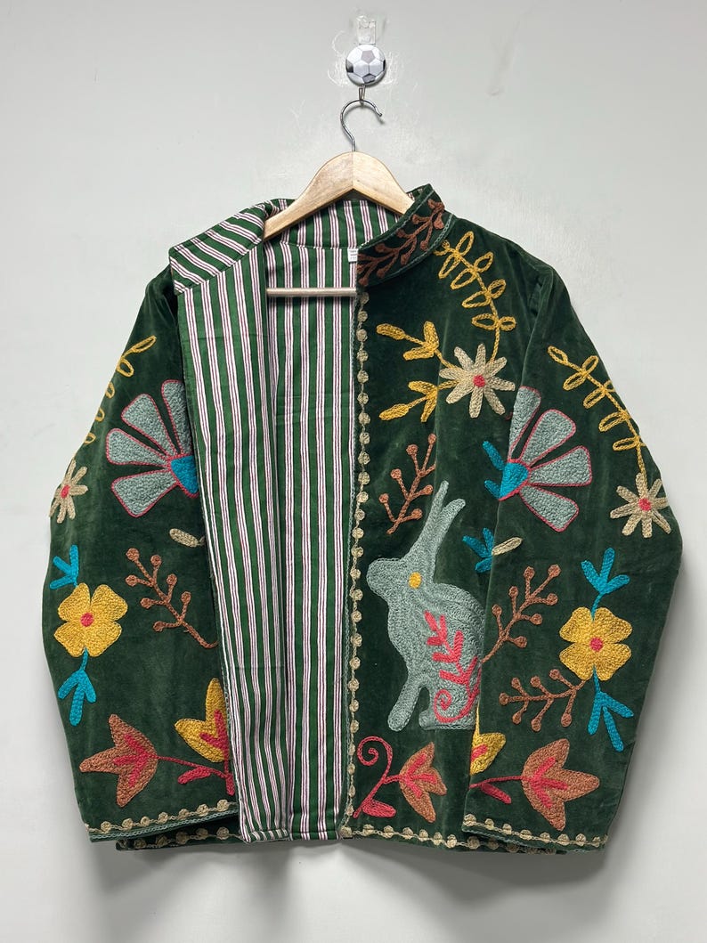 May include: A dark green velvet jacket featuring colorful embroidered floral patterns and a rabbit motif. The interior lining is made of white fabric with thin vertical green and red stripes. The jacket has long sleeves and a high collar.
