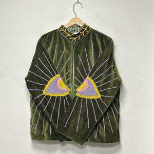 K&ouml;nnte beinhalten: Eine olivgr&uuml;ne Samtjacke mit gestickten Augenmotiven auf den Vorderteilen. Die Augen sind in lavendelfarbenem, rosa und gelbem Garn gehalten, mit silbernen Strahlen, die nach au&szlig;en verlaufen. Ein gelbes Rankenmuster ziert den Stehkragen.