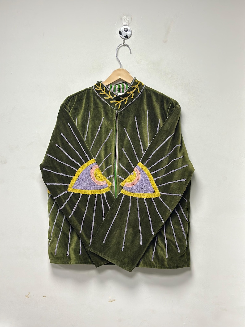 K&ouml;nnte beinhalten: Eine olivgr&uuml;ne Samtjacke mit gestickten Augenmotiven auf den Vorderteilen. Die Augen sind in lavendelfarbenem, rosa und gelbem Garn gehalten, mit silbernen Strahlen, die nach au&szlig;en verlaufen. Ein gelbes Rankenmuster ziert den Stehkragen.
