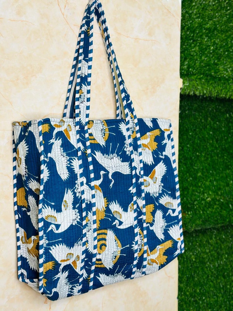 May include: A blue and white tote bag with a pattern of white cranes flying against a blue background. The bag has white and gold striped straps.