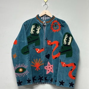 May include: Teal velvet jacket with embroidered whimsical designs. Features include green boots, orange snakes, a pink star, and an eye. The jacket has a zipper closure and is hanging on a wooden hanger.