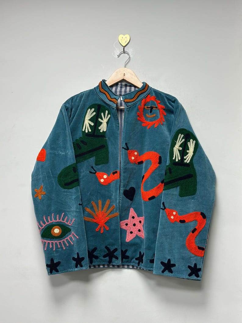May include: Teal velvet jacket with embroidered whimsical designs. Features include green boots, orange snakes, a pink star, and an eye. The jacket has a zipper closure and is hanging on a wooden hanger.