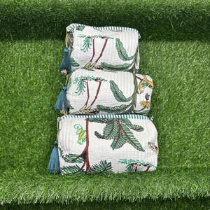May include: Three white quilted makeup bags with a tropical print featuring green leaves, red flowers, and yellow birds. The bags have blue tassels and a white and blue striped trim.