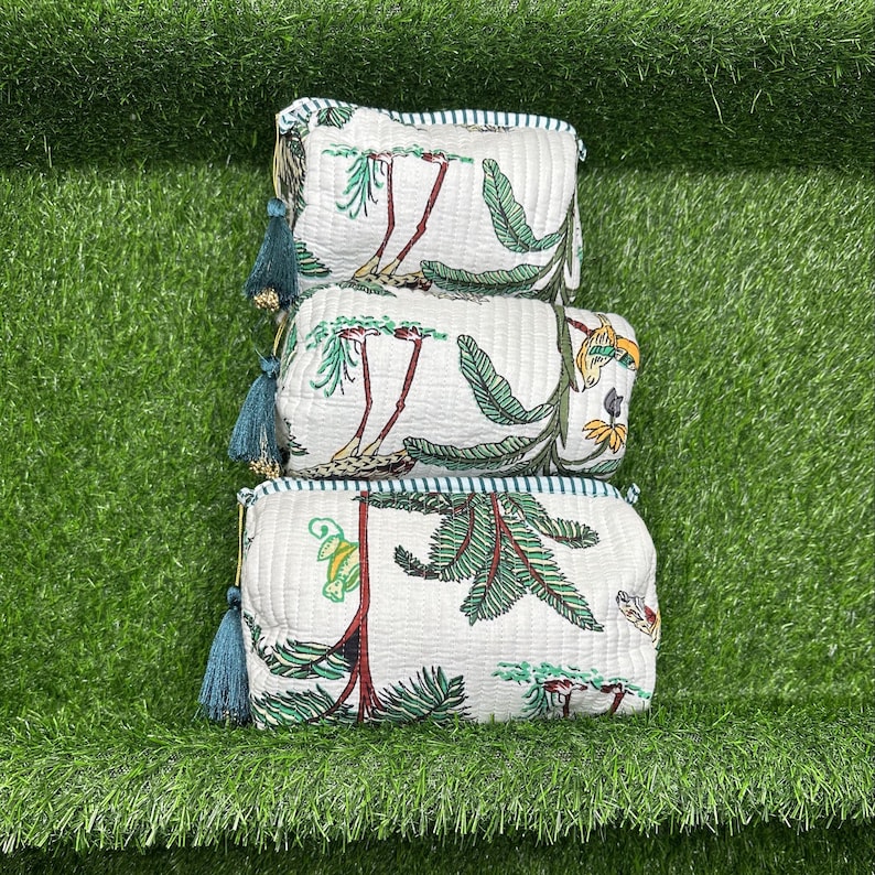 May include: Three white quilted makeup bags with a tropical print featuring green leaves, red flowers, and yellow birds. The bags have blue tassels and a white and blue striped trim.