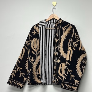 May include: A black jacket featuring long sleeves and an open front, adorned with intricate tan-coloured floral embroidery. The interior lining displays a vertical black and white striped pattern, visible along the front edges and collar.