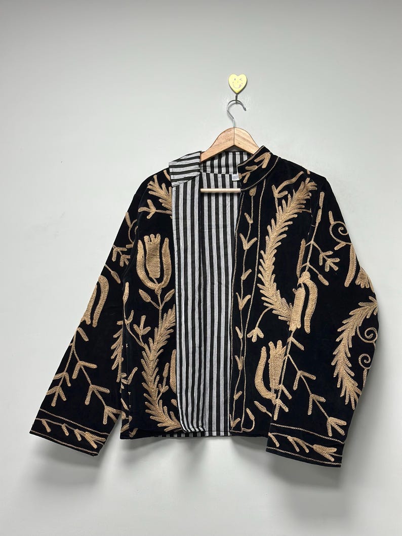 May include: A black jacket featuring long sleeves and an open front, adorned with intricate tan-coloured floral embroidery. The interior lining displays a vertical black and white striped pattern, visible along the front edges and collar.