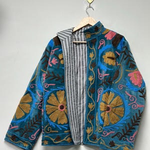 May include: A teal velvet jacket featuring intricate floral embroidery in shades of gold, pink, and brown. The jacket has a high collar, long sleeves, and an open front that reveals a black and white vertical striped lining on the interior.