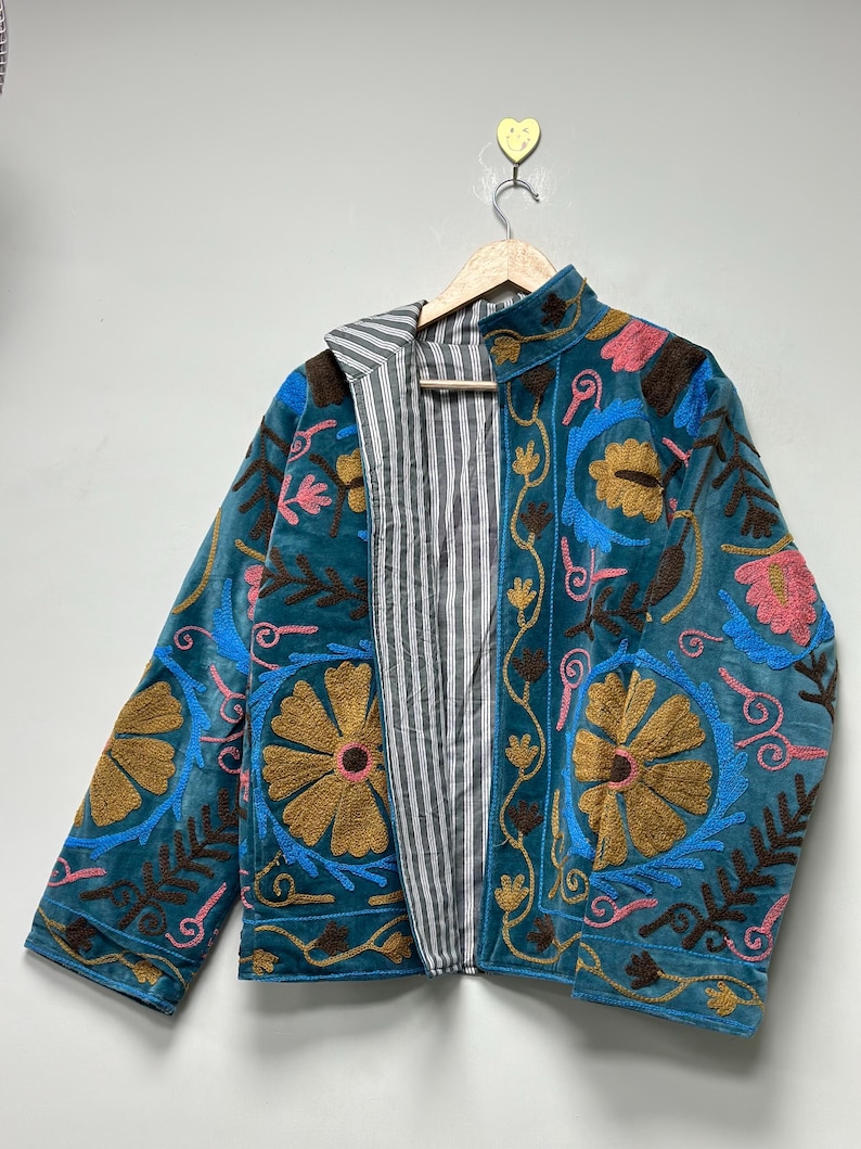 May include: A teal velvet jacket featuring intricate floral embroidery in shades of gold, pink, and brown. The jacket has a high collar, long sleeves, and an open front that reveals a black and white vertical striped lining on the interior.