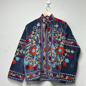 May include: A slate blue velvet jacket featuring intricate floral embroidery in shades of orange, magenta, and light blue. The garment has a high collar, long sleeves, and a front opening, with decorative stitching patterns throughout the fabric.