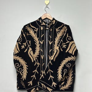 May include: A black jacket featuring intricate gold-coloured embroidery in a leaf and vine pattern. The garment has a high collar, long sleeves, and a front opening that reveals a black and white striped lining. It is displayed on a wooden hanger.
